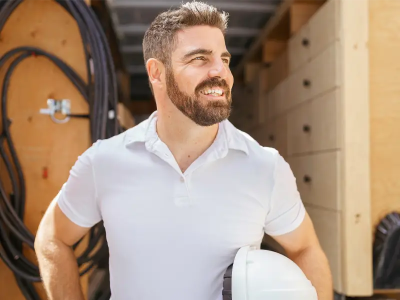 Bearded man in white shirt holding a white hard hat, standing in a workshop or storage area. damage restoration company